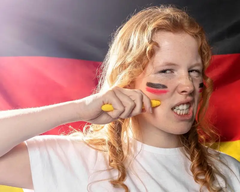Ragazza con capelli rossi e maglietta bianca ha una bandiera tedesca tatuata sul viso perché è fiera della cultura della Germania.