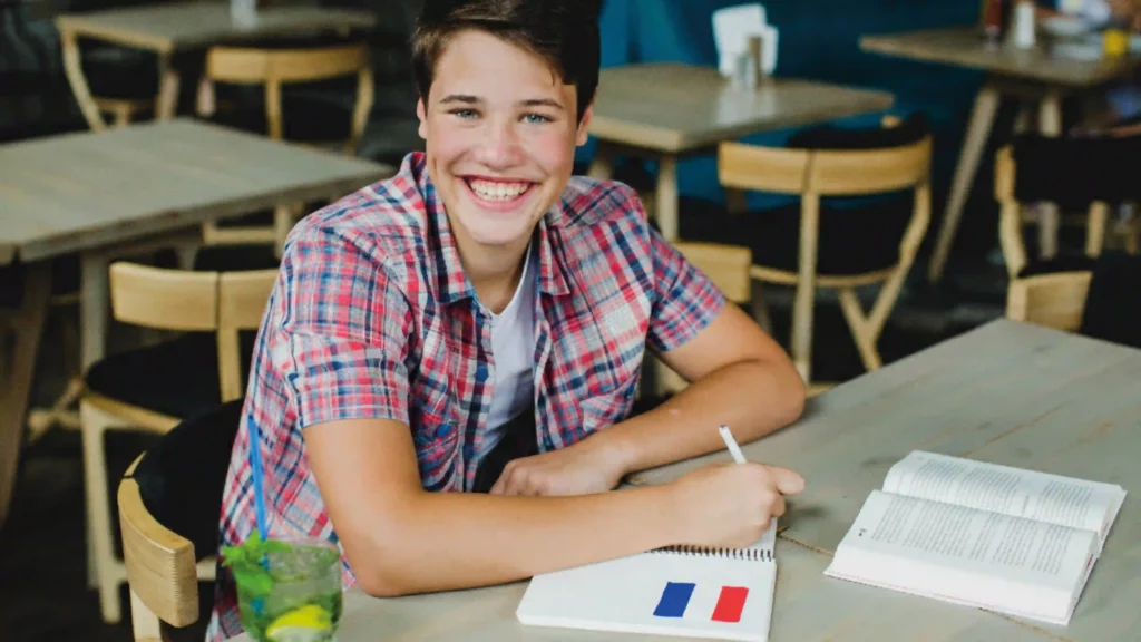 Giovane studente sorridente seduto al tavolo mentre scrive su un quaderno con la bandiera della Francia.