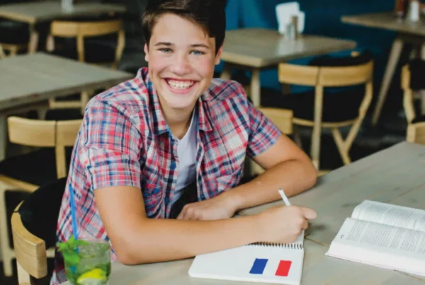 Giovane studente sorridente seduto al tavolo mentre scrive su un quaderno con la bandiera della Francia.