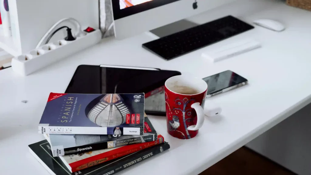 Libri per imparare lo spagnolo impilati su scrivania bianca accanto a tazza rossa e computer, pronti per lo studio
