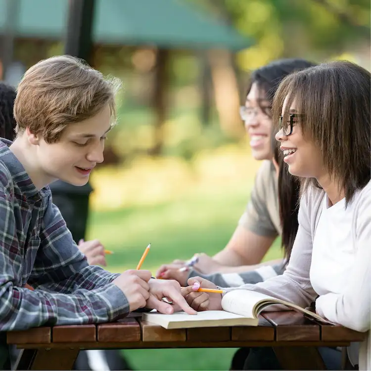 Studenti seduti a un tavolo di legno all'aperto mentre studiano e conversano, evidenziando l'importanza della pratica orale per imparare il francese.