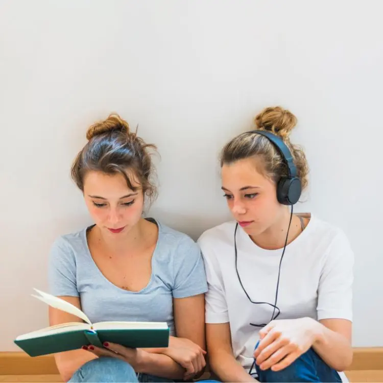 Due amiche adolescenti, una con le cuffie e la maglietta bianca, una con la maglia celeste, si siedono per terra e ripassano le regole per imparare insieme tedesco.