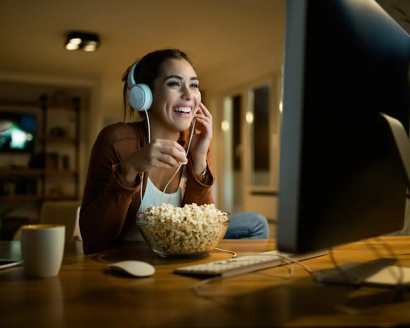 Ragazza che guarda serie TV in inglese al computer con cuffie e popcorn, divertita da una commedia britannica.
