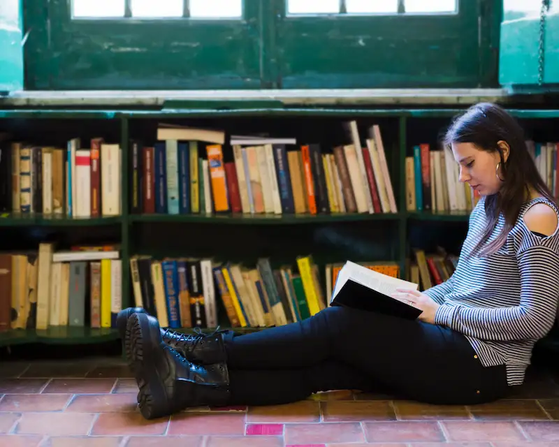 Ragazza seduta sul pavimento di una biblioteca che legge un libro in inglese