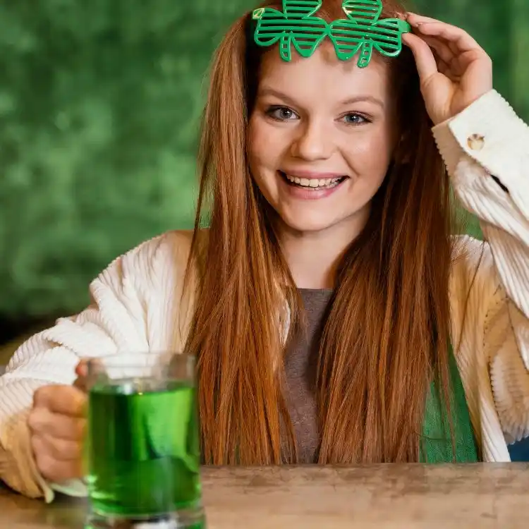 Ragazza con i capelli rossi beve birra verde e indossa occhiali con trifoglio. Una curiosità sull'irlanda è che questo simbolo ha origini celtiche.
