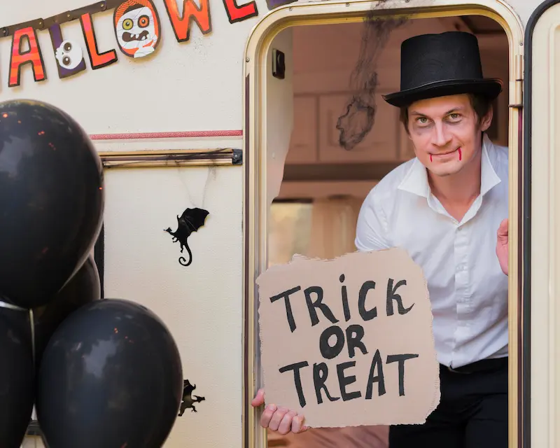 Un uomo travestito da vampiro mostra un cartello con la scritta Trick or Treat davanti a decorazioni a tema, illustrando le origini e i simboli di Halloween in inglese.