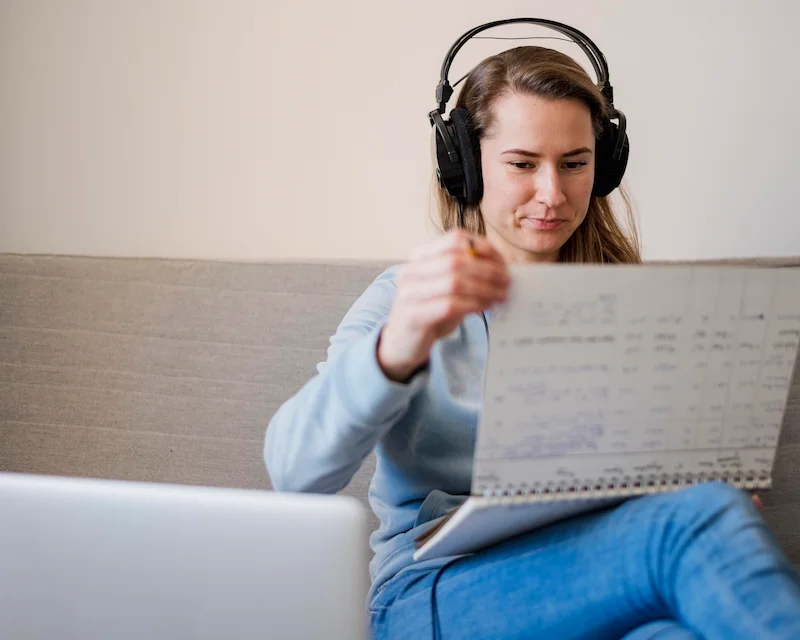 Donna con cuffie ascolta un podcast in inglese mentre prende appunti su un quaderno, laptop aperto davanti a sé"