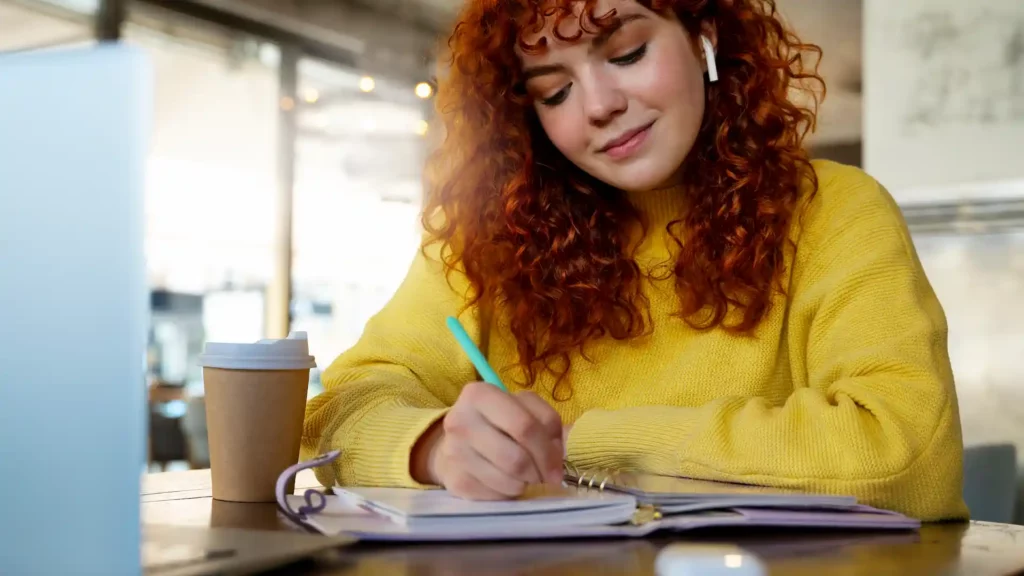 Ragazza con capelli ricci rossi scrive su un'agenda al tavolo di un bar, con laptop e caffè da asporto, mentre impara l'inglese velocemente
