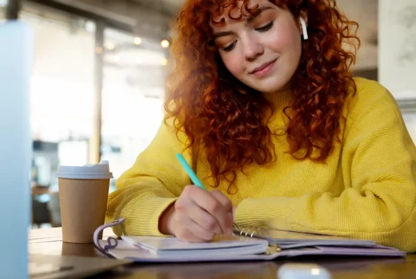 Ragazza con capelli ricci rossi scrive su un'agenda al tavolo di un bar, con laptop e caffè da asporto, mentre impara l'inglese velocemente