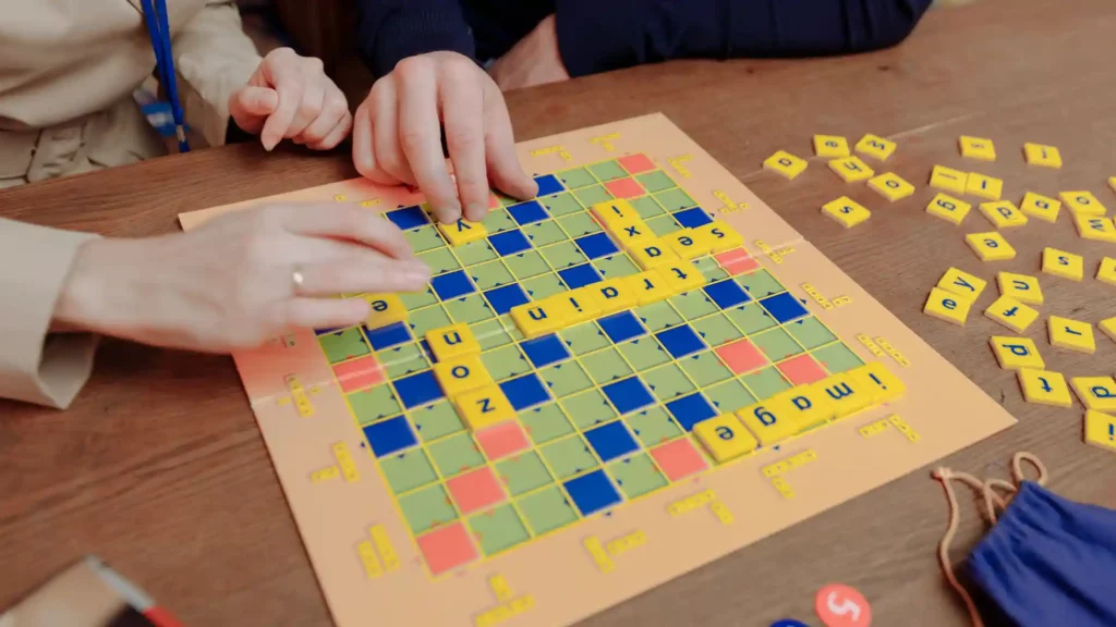 Due persone giocano a Scrabble su un tavolo in legno componendo parole in inglese con le tessere gialle.