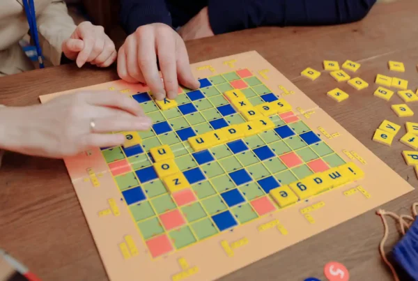 Due persone giocano a Scrabble su un tavolo in legno componendo parole in inglese con le tessere gialle.